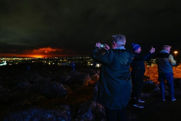 People take pictures with their pho<em></em>nes of a volcano eruption in the distance.